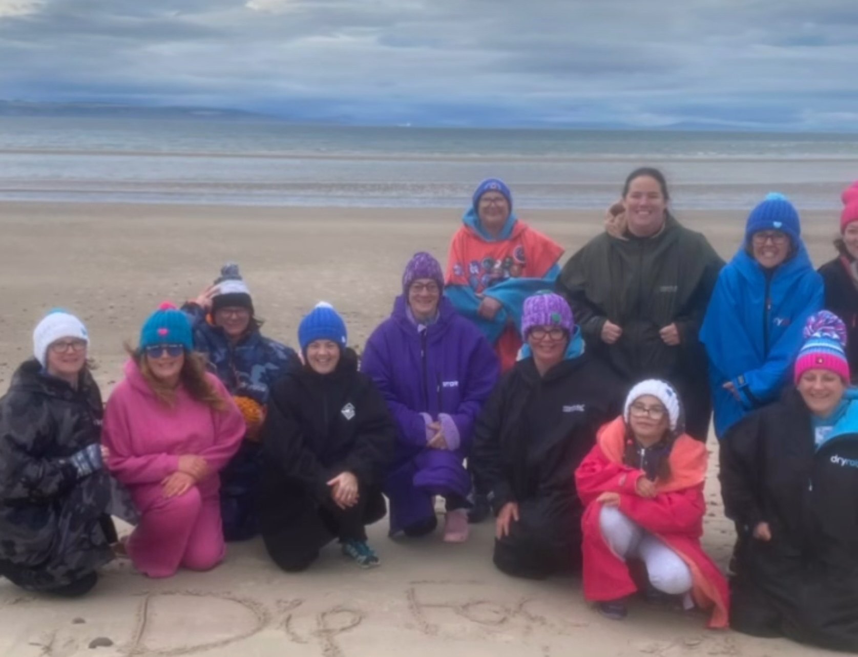 A group of cold water swimmers in coats and hats sitting together on a beach