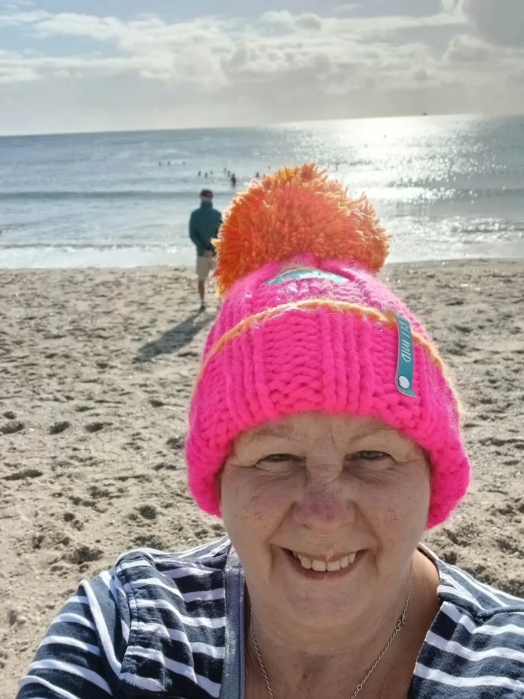 Ruth in a pink and orange bobble hat on the beach, with swimmers in the sea behind