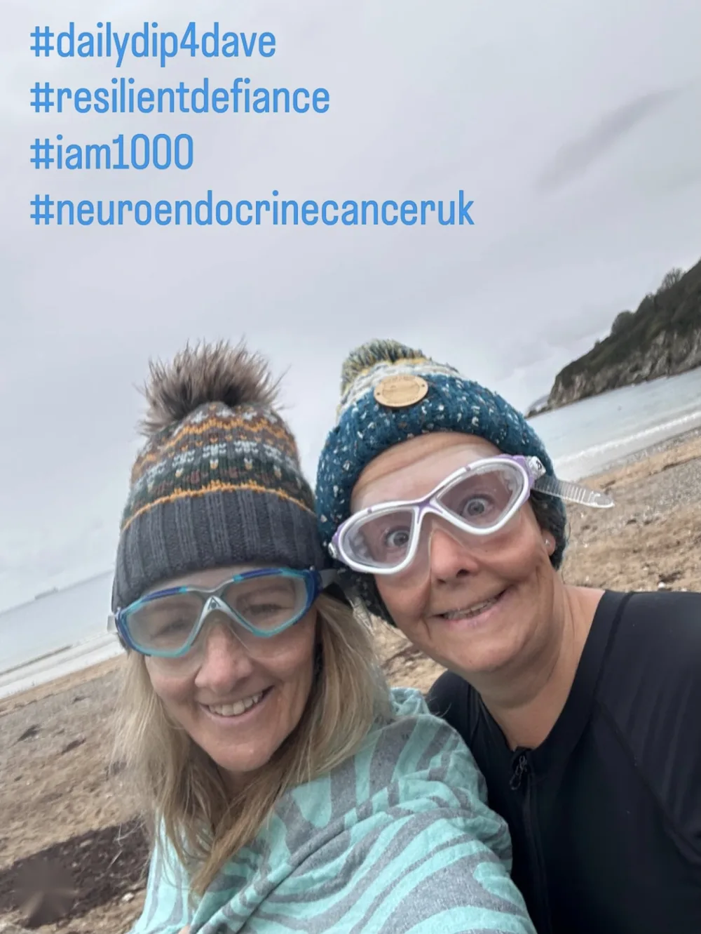 Lottie and MaryEllen in bobble hats and swimming goggles on Maenporth Beach, with the hashtags dailydip4dave, resilientdefiance, iam1000 and neuroendocrinecanceruk