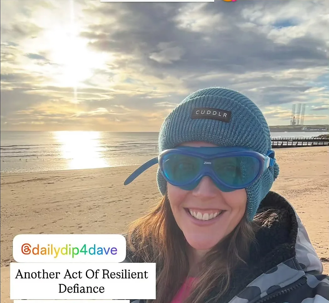 A smiling swimmer in goggles and a bobble hat on a beach at sunrise